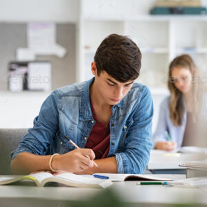 Focused young man taking notes from books for his study. College student sitting at desk with books for finding information in high school library. Guy studying in classroom and completing project with highschool classmates in background.